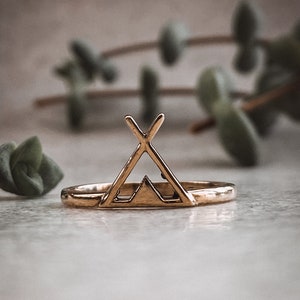 May include: A gold-toned ring with a geometric design of a tent with a mountain inside. The ring is on a light gray surface.