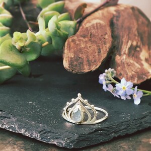 May include: A silver ring with a teardrop-shaped gemstone set in a crown-like design. The ring is on a dark gray slate surface.