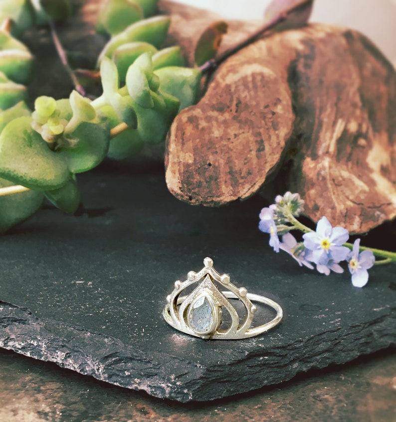 May include: A silver ring with a teardrop-shaped gemstone set in a crown-like design. The ring is on a dark gray slate surface.