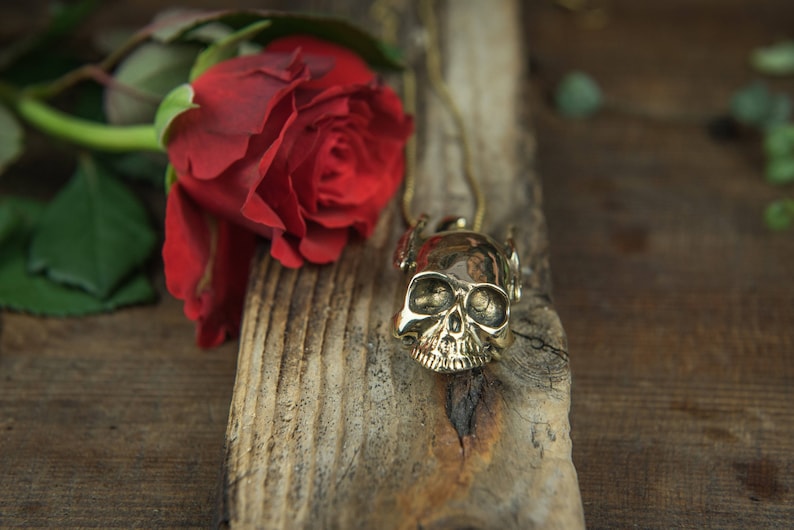 May include: A gold skull pendant necklace on a wooden surface with a red rose.