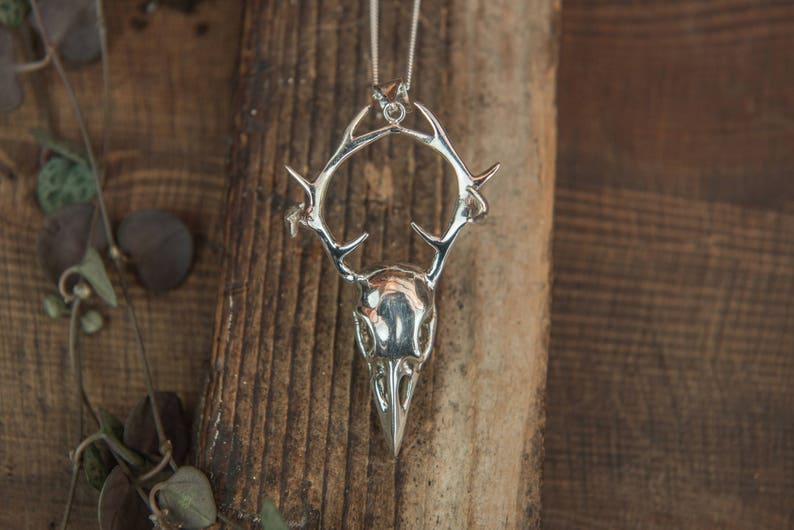 May include: A silver pendant necklace featuring a detailed skull with antlers. The pendant is on a wooden background.