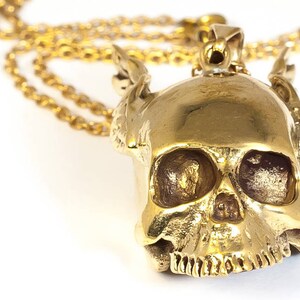 May include: A gold-colored skull pendant necklace with a detailed design. The skull has a textured surface and intricate features, including eye sockets, a nose, and a mouth.