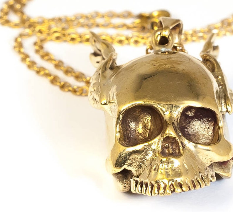 May include: A gold-colored skull pendant necklace with a detailed design. The skull has a textured surface and intricate features, including eye sockets, a nose, and a mouth.