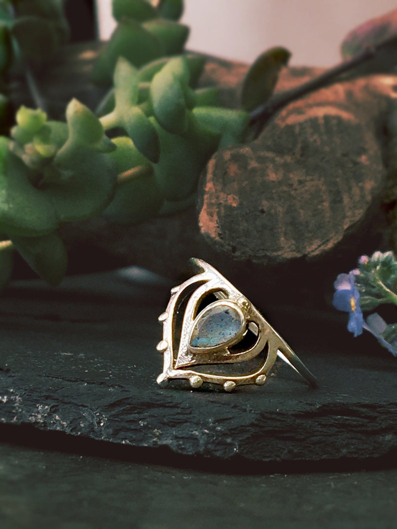 May include: A silver ring with a teardrop-shaped labradorite gemstone set in a delicate, ornate design. The ring is on a dark gray surface.