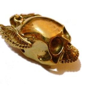 May include: A gold-colored skull pendant with wings. The skull is detailed with eye sockets, a nose, and teeth. The wings are spread out and have a detailed feather pattern.