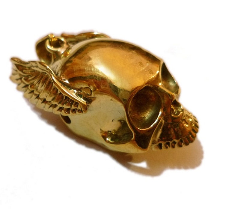 May include: A gold-colored skull pendant with wings. The skull is detailed with eye sockets, a nose, and teeth. The wings are spread out and have a detailed feather pattern.