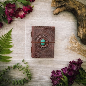 Könnte beinhalten: Ein braunes Lederjournal mit einem türkisfarbenen Steinbesatz und filigranen Blumenmustern. Das Journal ist umgeben von getrockneten Blumen, grünen Farnen und einem Stück Treibholz.