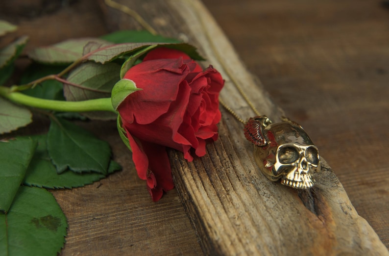 May include: A red rose with green leaves lies on a piece of weathered wood. A gold skull pendant with a red stone hangs from a chain.