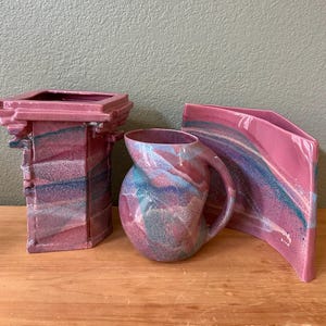 Puede incluir: Un conjunto de tres piezas de cerámica con un esmalte jaspeado en tonos rosa, azul y morado. La colección incluye un jarrón de borde cuadrado con detalles arquitectónicos, una jarra redondeada con asa y un jarrón curvo de lados planos.