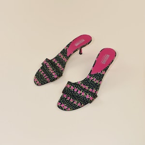 Op de afbeelding: Een paar zwarte en roze open teen hakken met bloemenprint. De hakken hebben een kleine stilettohak en een roze binnenzool met de merknaam "MOSCHINO" erop gedrukt. De schoenen staan tegen een neutrale achtergrond.