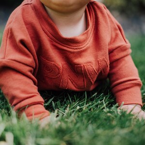 May include: A rust-colored baby sweatshirt with the letters "PNW" embroidered on the front. The child is crawling on green grass. The sweatshirt has long sleeves and a crew neck.