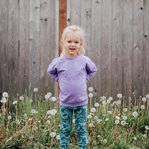 May include: A child in a lavender sweatshirt with the word "PINK" embroidered on the front, and patterned leggings with a teal and blue design. The child is standing in front of a wooden fence. The scene is outdoors.