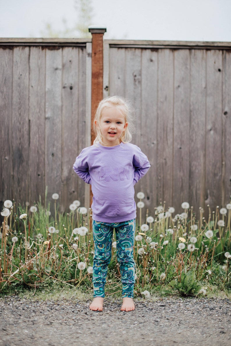 May include: A child in a lavender sweatshirt with the word "PINK" embroidered on the front, and patterned leggings with a teal and blue design. The child is standing in front of a wooden fence. The scene is outdoors.
