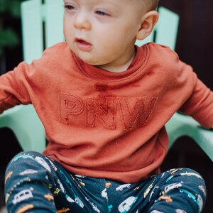 May include: A rust-colored sweatshirt with the letters "PNW" embroidered on the front. The child is wearing patterned pants with cars and trucks. The child is sitting in a light green chair.