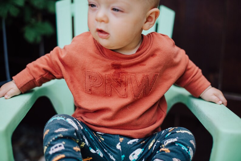 May include: A rust-colored sweatshirt with the letters "PNW" embroidered on the front. The child is wearing patterned pants with cars and trucks. The child is sitting in a light green chair.