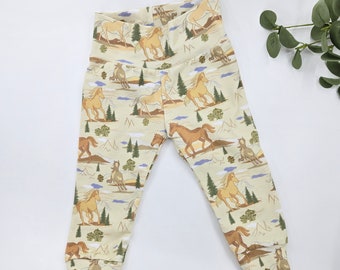 Puede incluir: Pantalones de bebé color crema con un estampado repetido de caballos corriendo, árboles y montañas. Los pantalones tienen una cintura alta y puños ajustados. Los caballos son en tonos marrones y beige.