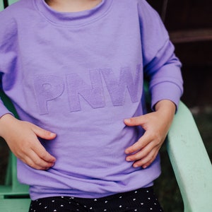 May include: A lilac crew neck sweatshirt with the letters "PNW" stitched across the chest. The sweatshirt has long sleeves and a relaxed fit. The child is wearing black and white polka dot trousers.
