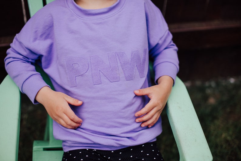 May include: A lilac crew neck sweatshirt with the letters "PNW" stitched across the chest. The sweatshirt has long sleeves and a relaxed fit. The child is wearing black and white polka dot trousers.