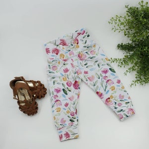 May include: White leggings with a floral print featuring pink, yellow, and blue watercolor flowers. The leggings have a cuffed hem.