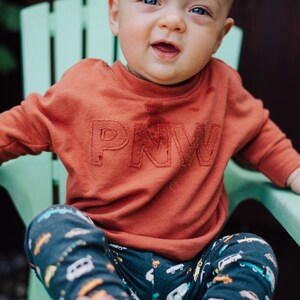 May include: A child wearing a rust-colored sweatshirt with the letters "PNW" embroidered on the front. The child is sitting in a light green chair and wearing patterned pants with cars and trucks.