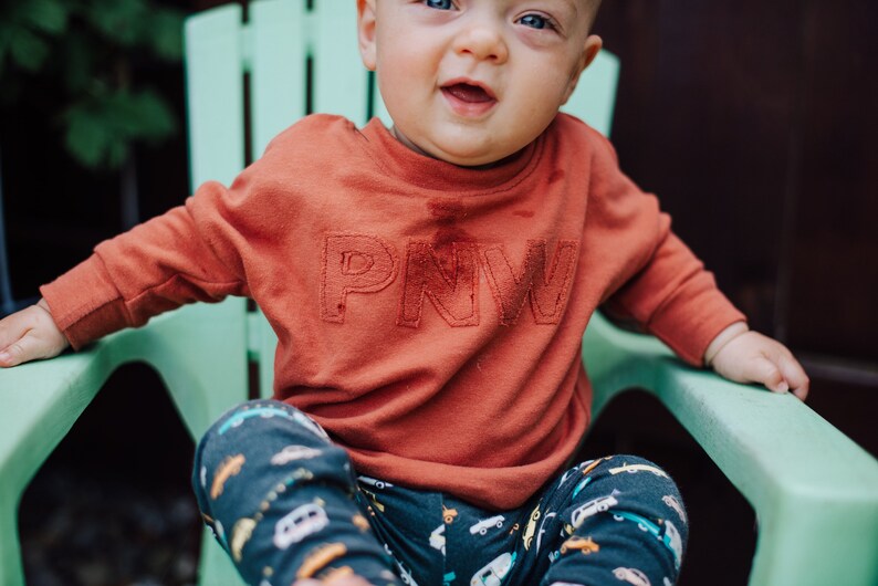 May include: A child wearing a rust-colored sweatshirt with the letters "PNW" embroidered on the front. The child is sitting in a light green chair and wearing patterned pants with cars and trucks.
