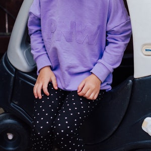 May include: A lilac sweatshirt with the letters "PNW" embroidered on the front. The sweatshirt is paired with black leggings patterned with white hearts. The child is sitting next to a black toy car.