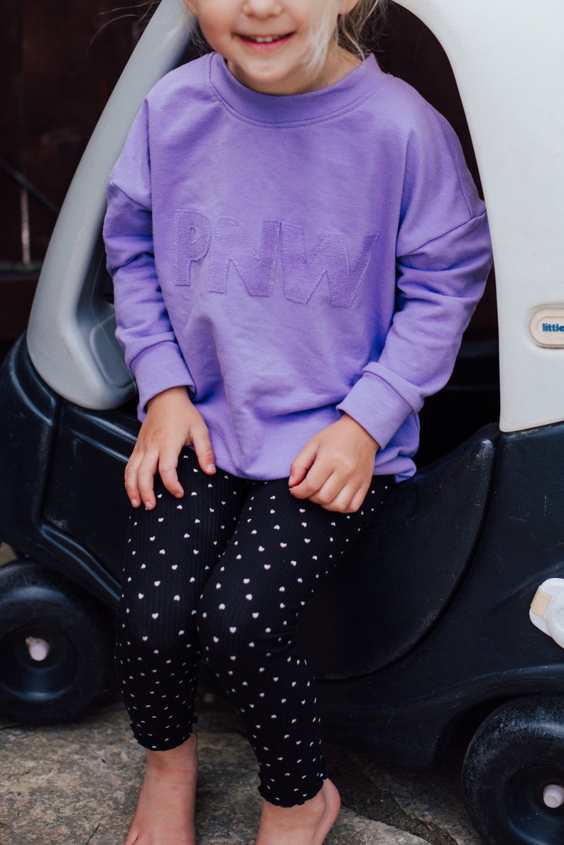 May include: A lilac sweatshirt with the letters "PNW" embroidered on the front. The sweatshirt is paired with black leggings patterned with white hearts. The child is sitting next to a black toy car.
