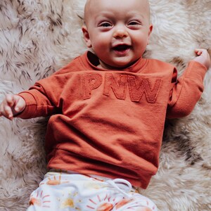 May include: A smiling baby wearing an orange sweatshirt with "PNW" stitched across the chest and white pants with a sun pattern. The baby is lying on a fluffy, white and brown rug.