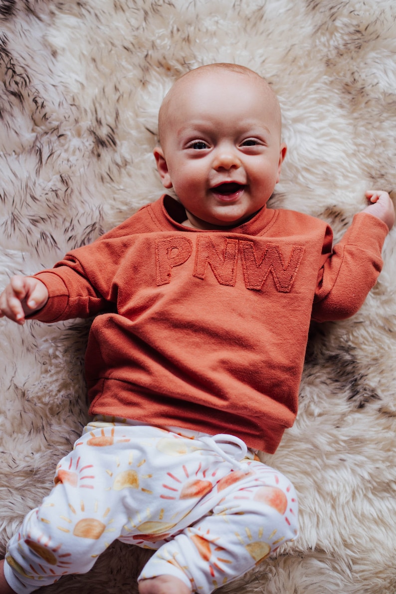 May include: A smiling baby wearing an orange sweatshirt with "PNW" stitched across the chest and white pants with a sun pattern. The baby is lying on a fluffy, white and brown rug.