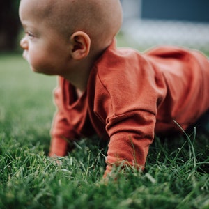 May include: A baby wearing an orange long-sleeved shirt and blue pants crawls on green grass. The baby is bald and facing left, with a focused expression. The image is taken outdoors in natural light.
