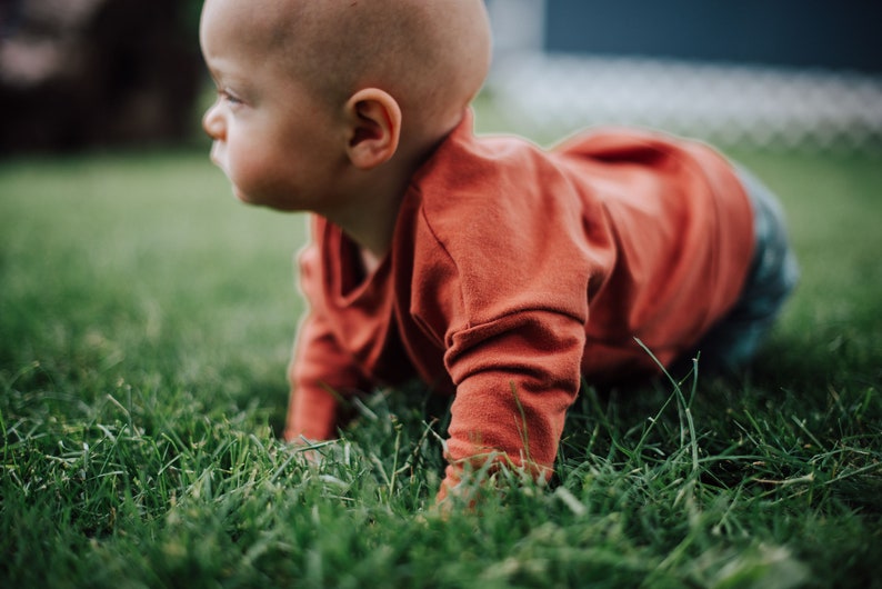 May include: A baby wearing an orange long-sleeved shirt and blue pants crawls on green grass. The baby is bald and facing left, with a focused expression. The image is taken outdoors in natural light.