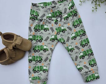 Puede incluir: Pantalones de bebé grises con un estampado repetido de camiones de basura verdes, conos de tráfico naranjas y formas negras con forma de hoja. Los pantalones tienen un puño ajustado en los tobillos. Un par de zapatos de bebé de estilo mocasín color canela están a la izquierda.