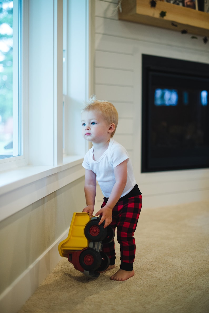 Puede incluir: Un ni&ntilde;o peque&ntilde;o con una camisa blanca y pantalones de cuadros rojos y negros sostiene un cami&oacute;n de volteo de juguete amarillo. El ni&ntilde;o est&aacute; mirando por una ventana.