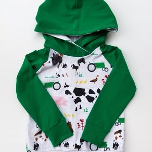 May include: A long-sleeve hooded sweatshirt featuring a solid green hood and sleeves with a white body. The body displays a farm-themed print with cows, horses, tractors, ducks, and red weather vanes on a white background with green grass.