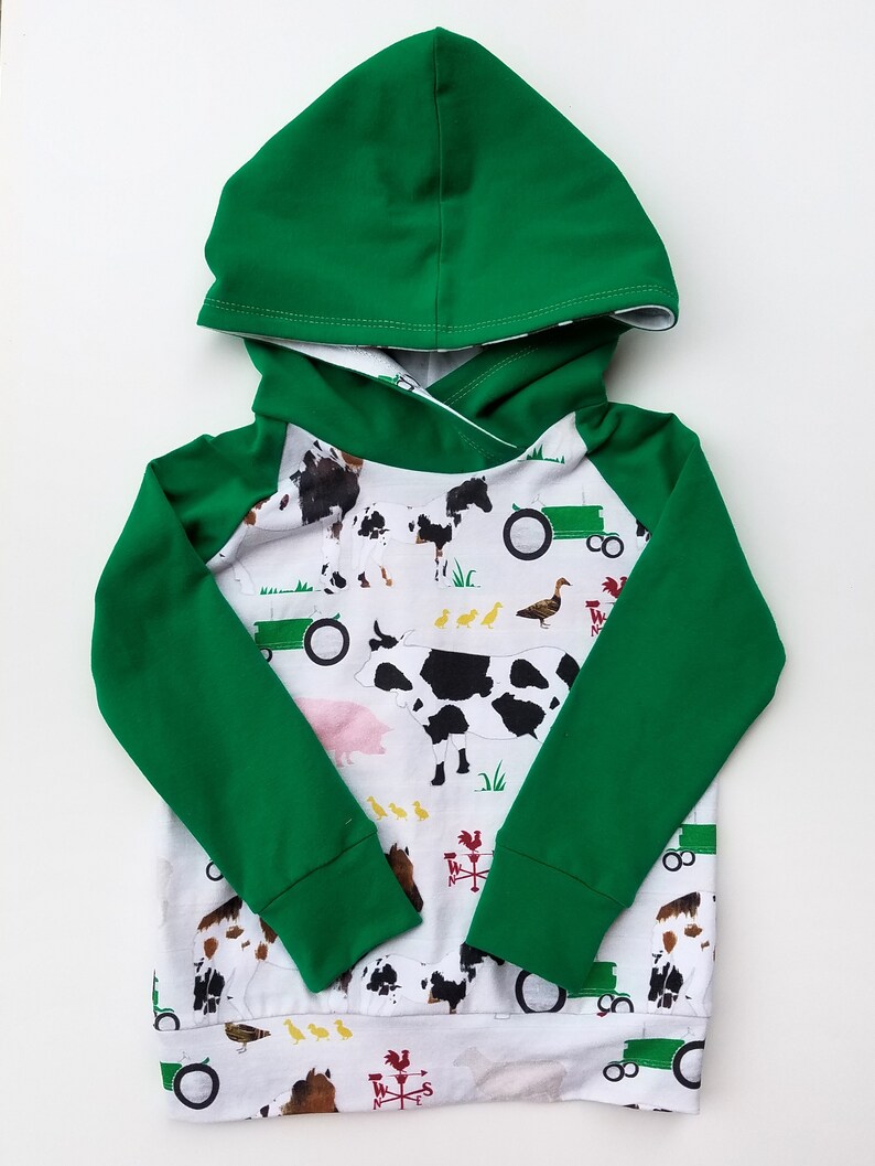May include: A long-sleeve hooded sweatshirt featuring a solid green hood and sleeves with a white body. The body displays a farm-themed print with cows, horses, tractors, ducks, and red weather vanes on a white background with green grass.