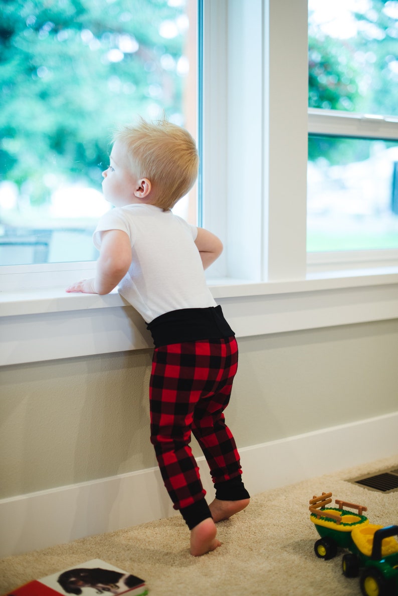 Puede incluir: Un ni&ntilde;o peque&ntilde;o con una camisa blanca y pantalones de cuadros rojos y negros se para junto a una ventana mirando hacia afuera.