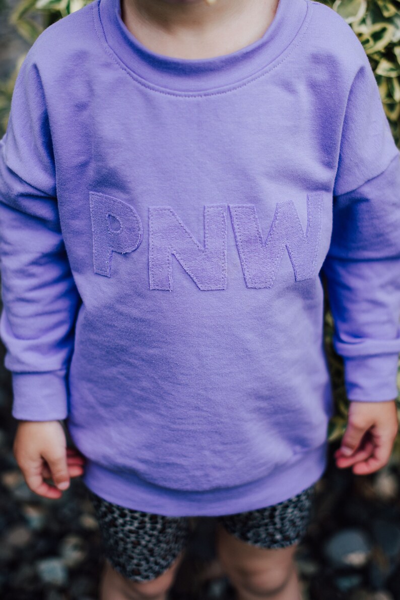 May include: A lilac sweatshirt featuring the embroidered letters "PNW". The long-sleeved top is made of a soft material and is worn by a child. The child is also wearing patterned shorts.