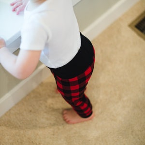 Puede incluir: Un ni&ntilde;o peque&ntilde;o con una camisa blanca y pantalones de ch&aacute;ndal de cuadros rojos y negros. El ni&ntilde;o est&aacute; de pie sobre una alfombra beige.
