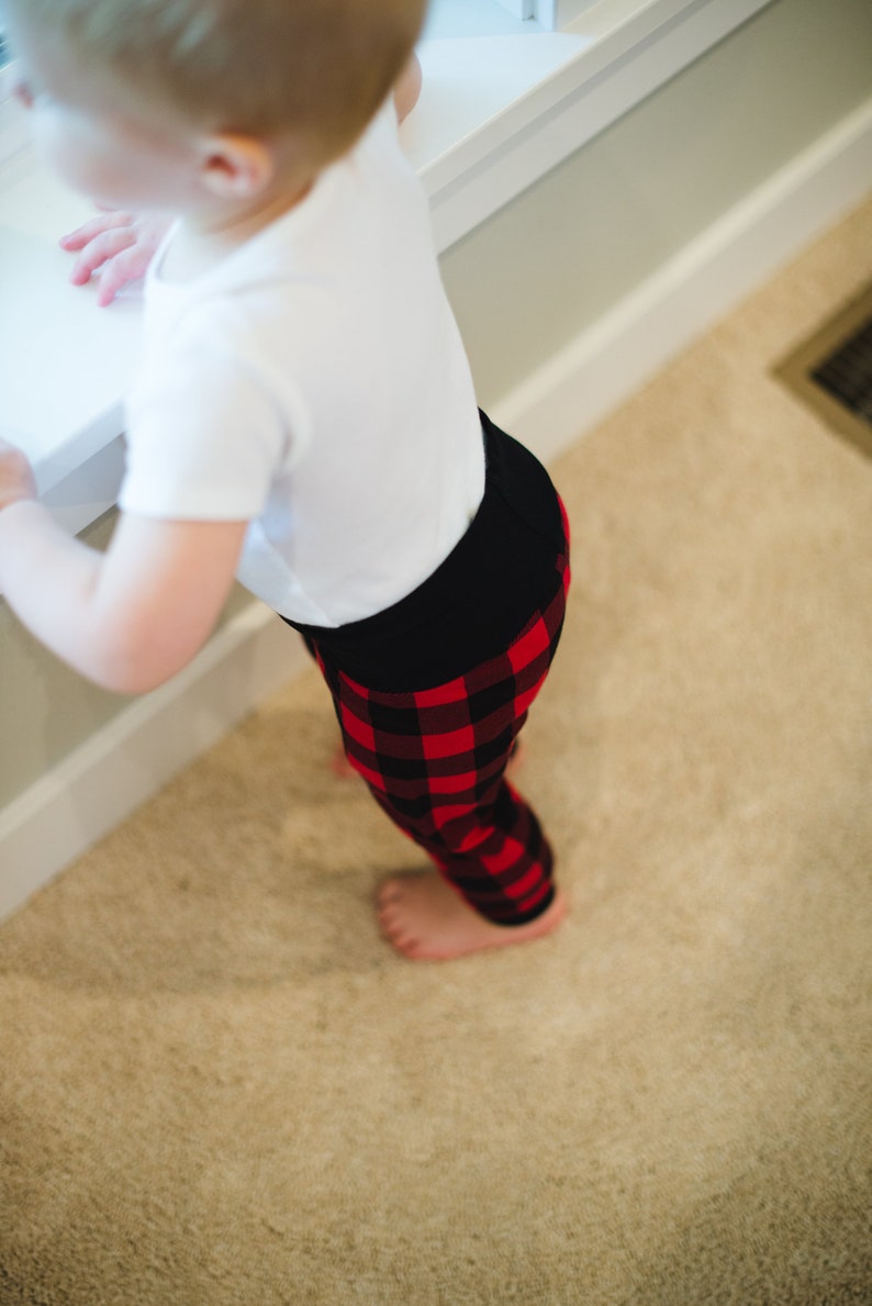 Puede incluir: Un ni&ntilde;o peque&ntilde;o con una camisa blanca y pantalones de ch&aacute;ndal de cuadros rojos y negros. El ni&ntilde;o est&aacute; de pie sobre una alfombra beige.