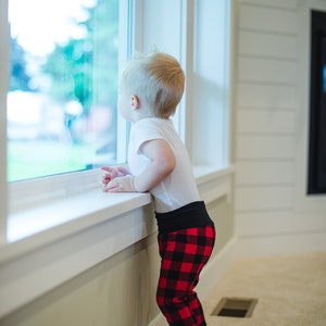 Puede incluir: Un ni&ntilde;o peque&ntilde;o con una camisa blanca y pantalones de cuadros rojos y negros se para junto a una ventana mirando hacia afuera.