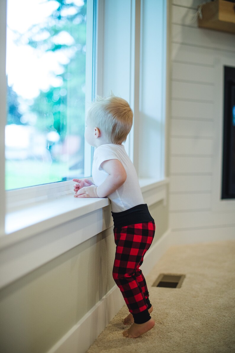 Puede incluir: Un ni&ntilde;o peque&ntilde;o con una camisa blanca y pantalones de cuadros rojos y negros se para junto a una ventana mirando hacia afuera.
