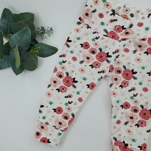 May include: White baby leggings with a floral print featuring pink and red flowers, green leaves, and black stems on a white background.