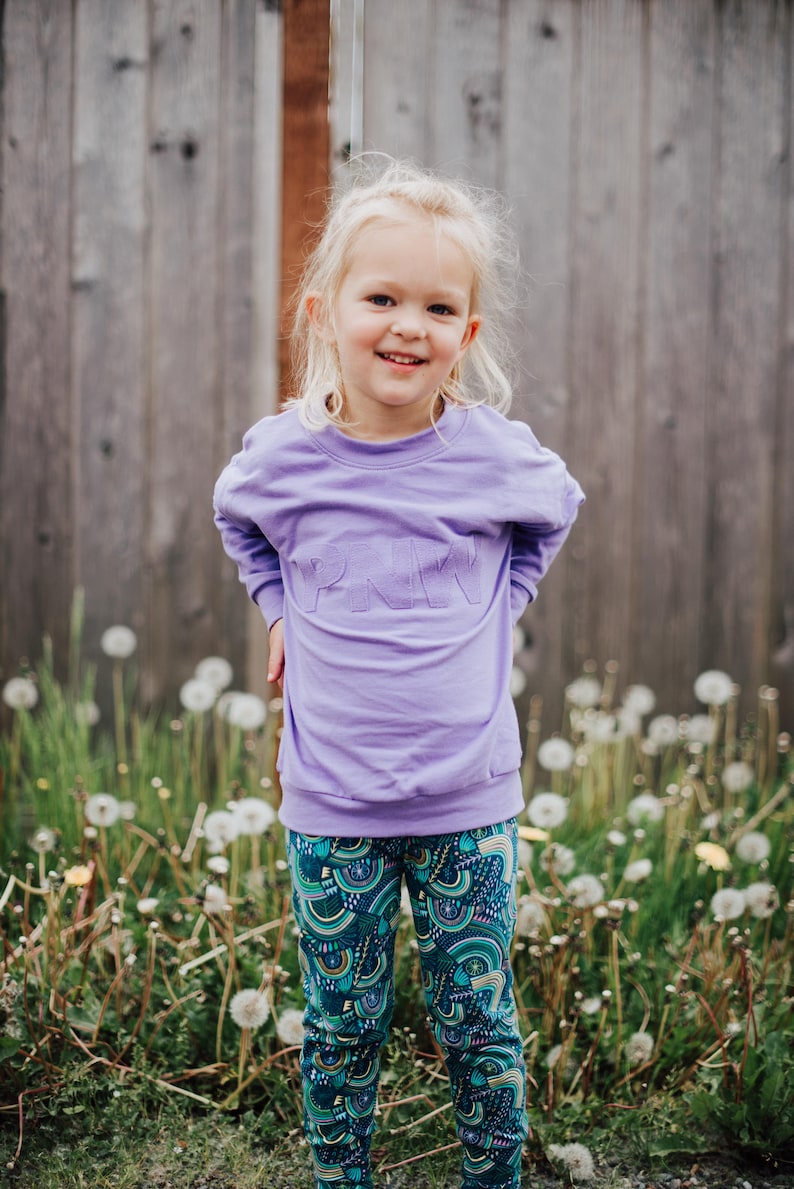 May include: A child in a lavender sweatshirt with "PNW" lettering and patterned leggings. The leggings feature a teal background with a colourful, abstract design. The child smiles in front of a wooden fence.