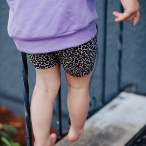 May include: A child wearing a lilac sweatshirt and leopard print shorts. The shorts feature a black and tan repeating animal print pattern. The child is standing on a concrete surface.
