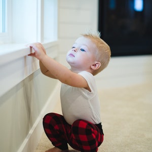Puede incluir: Un ni&ntilde;o peque&ntilde;o con una camisa blanca y pantalones de cuadros rojos y negros est&aacute; sentado en el suelo mirando hacia arriba. El ni&ntilde;o est&aacute; sentado cerca de una ventana y tiene la mano en el alf&eacute;izar de la ventana.
