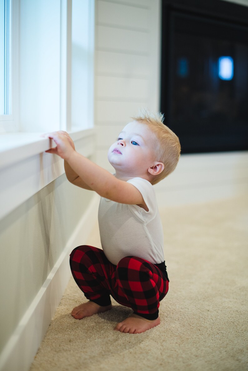 Puede incluir: Un ni&ntilde;o peque&ntilde;o con una camisa blanca y pantalones de cuadros rojos y negros est&aacute; sentado en el suelo mirando hacia arriba. El ni&ntilde;o est&aacute; sentado cerca de una ventana y tiene la mano en el alf&eacute;izar de la ventana.