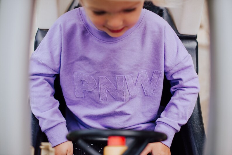 May include: A lilac crew neck sweatshirt with the letters "PNW" stitched across the chest. The sweatshirt is a solid colour and has long sleeves. The image is of a child wearing the sweatshirt.