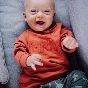 May include: A smiling baby wearing an orange sweatshirt with the letters "PNW" and patterned pants. The baby is lying on a gray upholstered surface.
