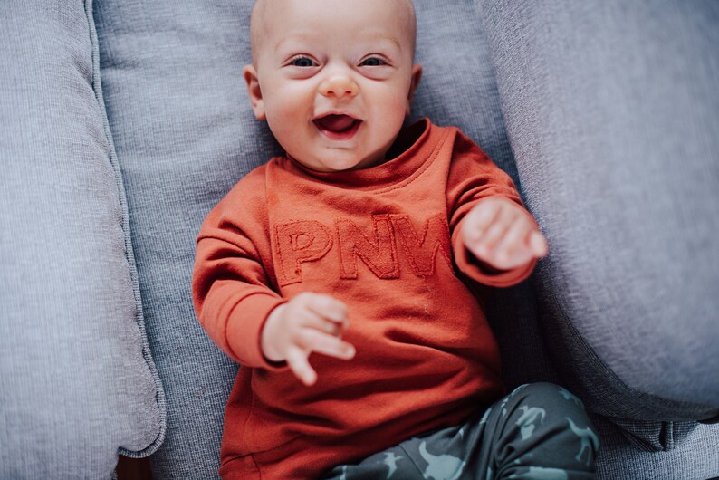 May include: A smiling baby wearing an orange sweatshirt with the letters "PNW" and patterned pants. The baby is lying on a gray upholstered surface.