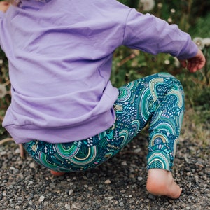 May include: A child in a lavender sweatshirt and patterned leggings. The leggings have a teal background with a repeating design of green, yellow, and white arches and lines. The child is squatting on a gravel surface.